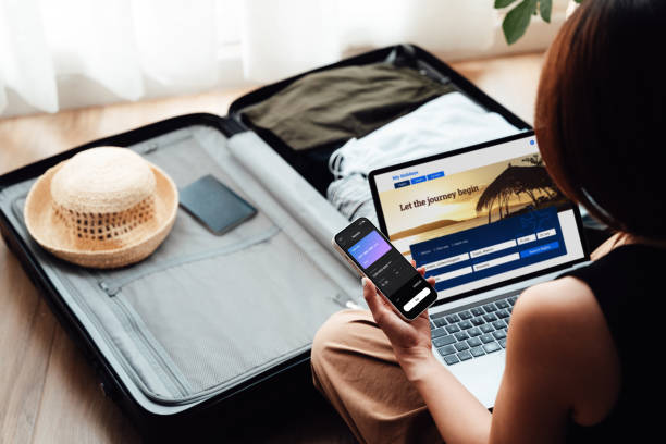 over the shoulder view of young woman booking a flight via laptop and smartphone for vacation - travel stock pictures, royalty-free photos & images