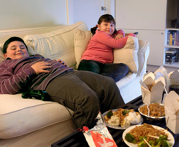 overweight brother and sister sitting on a sofa eating takeaway food and watching the tv - junk food stock pictures, royalty-free photos & images
