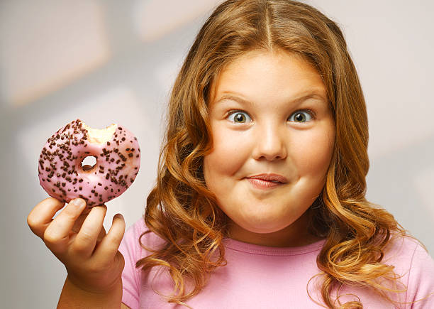 overweight girl (8-9) eating doughnut, smiling, portrait, close-up - junk food stock pictures, royalty-free photos & images