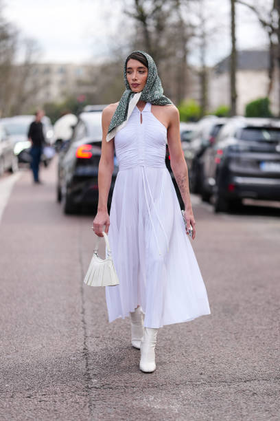 Penelope Guerrero wears black sunglasses, dark green pattern/print head scarf, white halter pleated maxi dress, cream leather bag, white heeled boots...