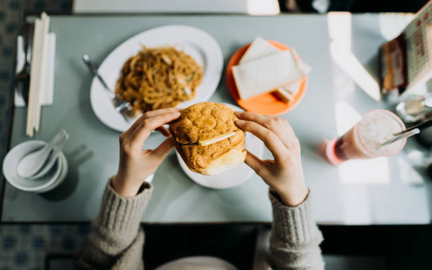 personal perspective of woman eating hong kong style local foods, sandwich, buttered pineapple bun, fried noodles and iced red bean drink in traditional restaurant - junk food stock pictures, royalty-free photos & images