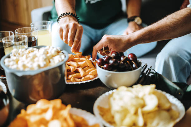 picture of hands of unrecognizable young people sitting on couch reaching for plates with yummy snacks - junk food stock pictures, royalty-free photos & images