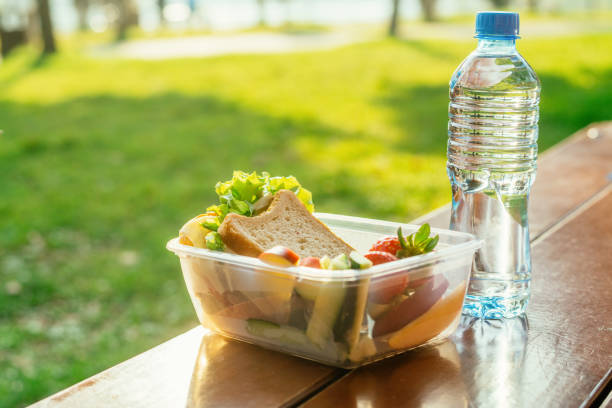 plastic container with sandwich, vegetables and fruits and a bottle of water on a table in a park in sunset - junk food stock pictures, royalty-free photos & images