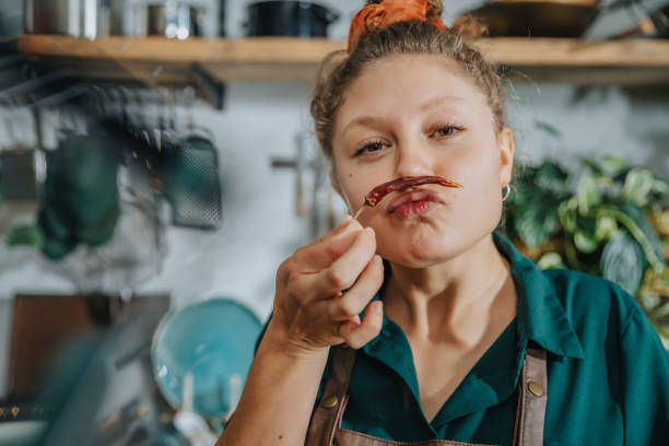 playful chef making mustache with dry chili pepper while standing in kitchen - food stock pictures, royalty-free photos & images