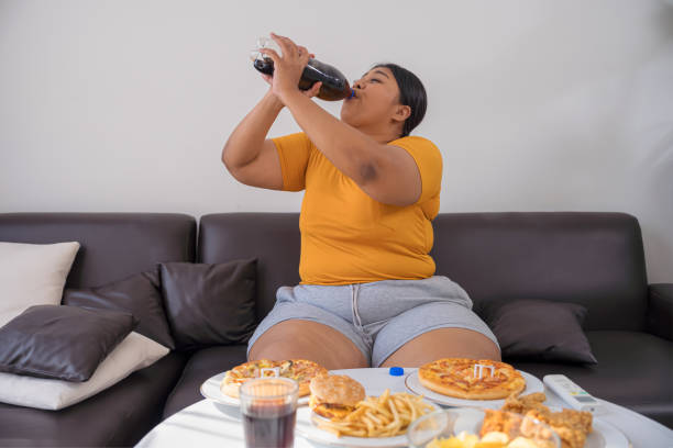 plus size woman drinking cola while eating junk food on sofa. - junk food stock pictures, royalty-free photos & images