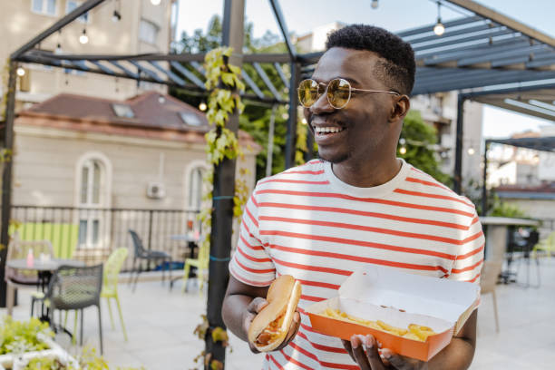 portrait of a young african-american man is eating hot dog and smiling - junk food stock pictures, royalty-free photos & images