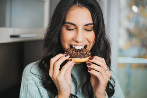 portrait of a young woman eating a donut in the kitchen - junk food stock pictures, royalty-free photos & images