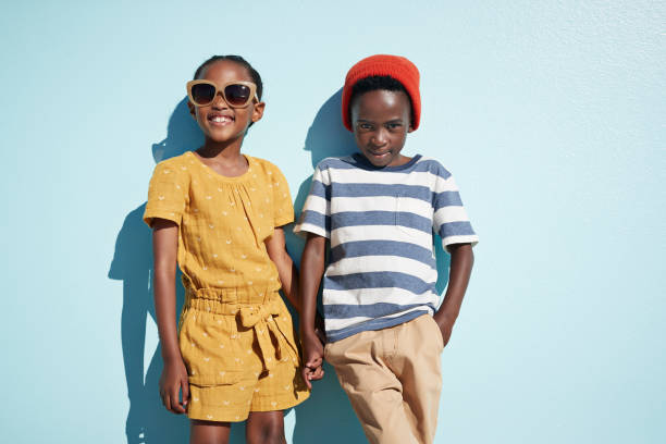portrait of boy & girl holding hands and looking in camera, on blue backdrop in summer - fashion photos et images de collection
