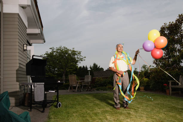 portrait of elderly man wearing birthday hat and streamers and holding balloons - garden decoration stock pictures, royalty-free photos & images
