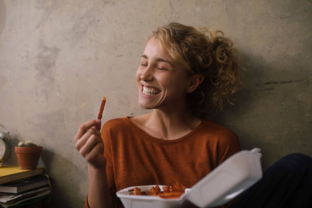 portrait of grinning young woman eating french fries at home - junk food stock pictures, royalty-free photos & images