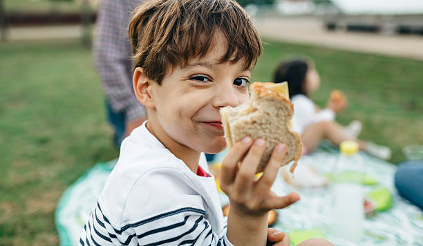 portrait of smiling boy holding sandwich with his family in background - food stock pictures, royalty-free photos & images