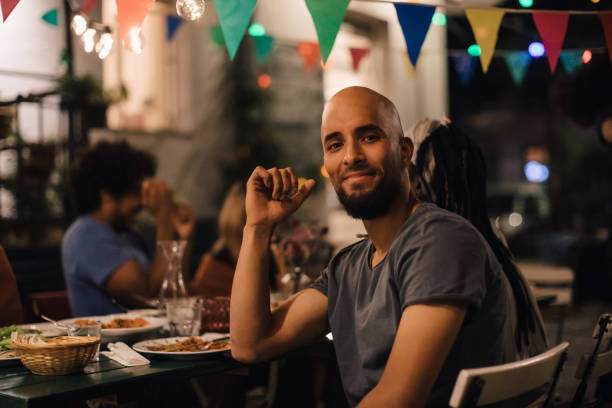 portrait of smiling young man with shaved head sitting at table during dinner party in backyard - garden decoration stock pictures, royalty-free photos & images
