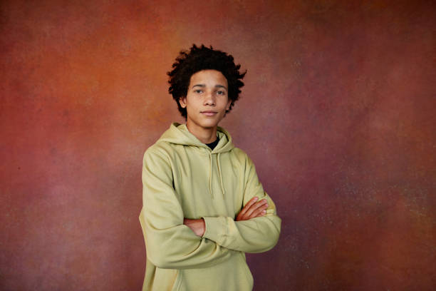 portrait of teenage boy in hooded shirt with arms crossed standing against brown background - fashion photos et images de collection