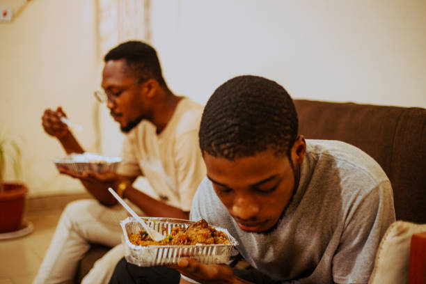 portrait of two men eating takeout food on sofa at home,tema,ghana - junk food stock pictures, royalty-free photos & images