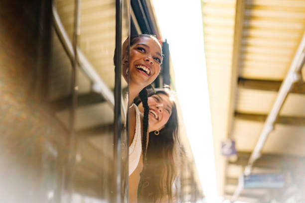 portrait shot of two young women looking out of an open door of a train on a platform and smiling - travel stock pictures, royalty-free photos & images