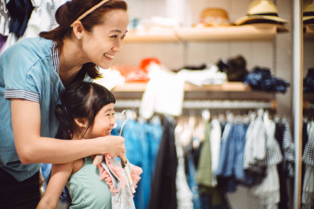 pretty young mom trying on clothes with her lovely little daughter in a fashion boutique joyfully. - fashion photos et images de collection