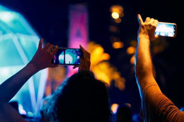 rear view of a long haired woman standing with a group of people at a concert recording with her phone - concert stock pictures, royalty-free photos & images
