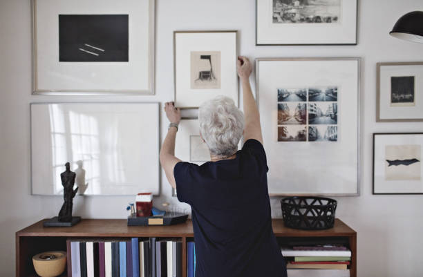 rear view of retired senior woman adjusting picture frame on wall over bookshelf at home - home decoration stock pictures, royalty-free photos & images