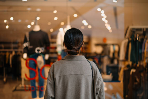 rear view of young asian woman standing outside a boutique looking at clothings through shop window while shopping in shopping mall. window shopping. sale season. seasonal shopping and consumerism concept - fashion stock pict