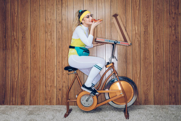 retro style exercise bike woman eighties era eating pizza - junk food stock pictures, royalty-free photos & images