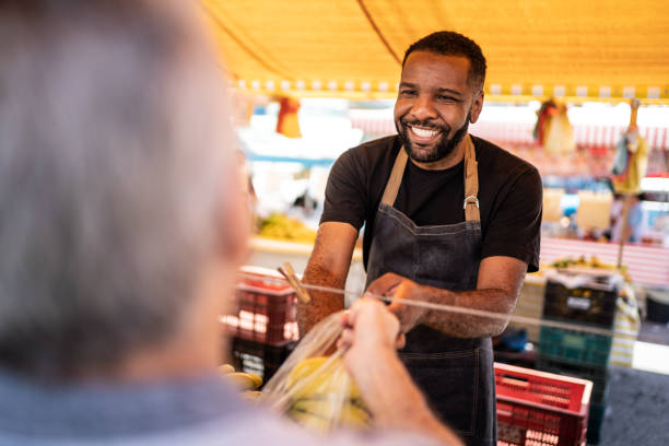 salesman helping the his customer putting the bananas in a plastic bag on a street market - food stock pictures, royalty-free photos & images