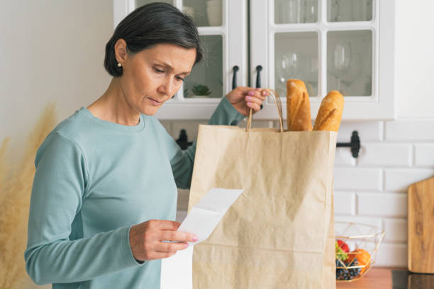 senior adult female worried while looking at receipt from grocery store. rise in price of products concept. buying consumer goods during inflation. facing economic downturn. kitchen unit and paper bag on background, horizonta