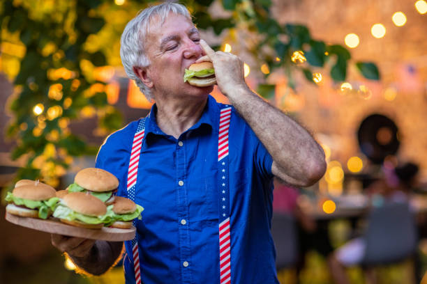 senior man eating a hamburger on celebration of american holiday barbecue party - junk food stock pictures, royalty-free photos & images