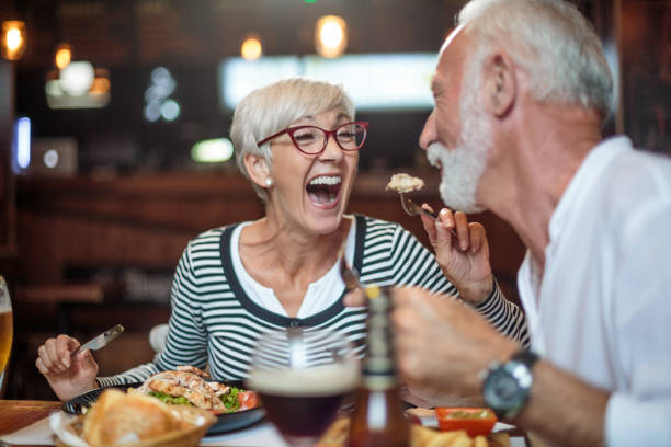 senior woman laughing while feeding her male partner in the restaurant - food stock pictures, royalty-free photos & images
