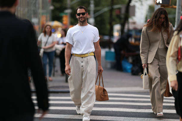 Simon Porte Jacquemus is seen wearing black Wayfarer sunglasses from Ray-Ban, a fine gold necklace with a rectangular pendant, an oversized white...
