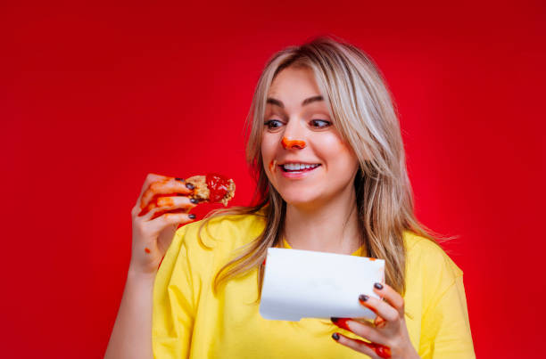 smiling blonde woman with red nose eating fried chicken wings in sauce in red studio - junk food stock pictures, royalty-free photos & images