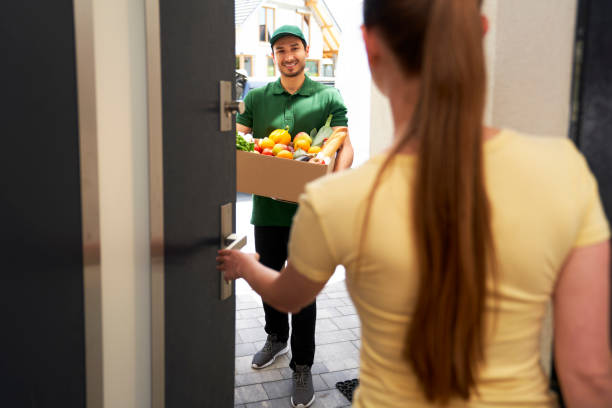smiling deliveryman holding vegetable box while standing at entrance - food stockfoto's en -beelden