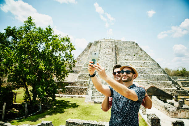 smiling gay couple taking selfie with smartphone while exploring mayapan ruins during vacation - travel stock-fotos und bilder