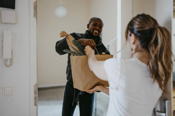 smiling man giving paper bag of groceries to woman standing at doorway - food stock pictures, royalty-free photos & images