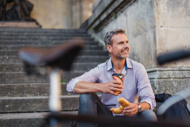 smiling man sitting on stairs with croissant and takeaway coffee in the city - junk food stock pictures, royalty-free photos & images