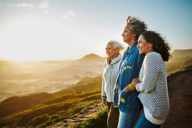 smiling mature friends standing arm in arm by a scenic coast during a road trip - travel stock pictures, royalty-free photos & images