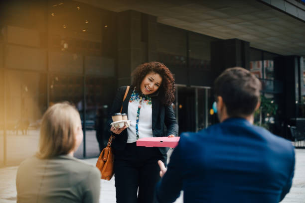 smiling mid adult businesswoman talking on mobile phone while giving food and drinks to colleagues against cafe - junk food stock pictures, royalty-free photos & images