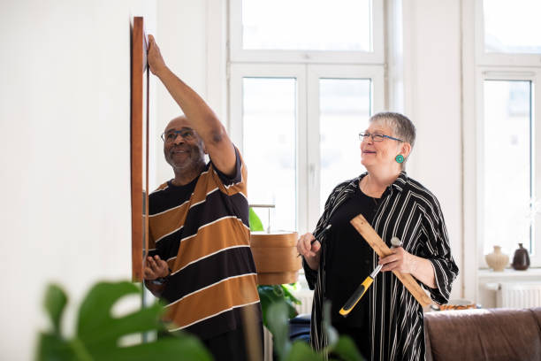 smiling senior couple hanging painting on wall at home - home decoration stockfoto's en -beelden