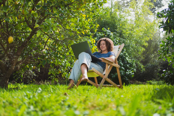smiling woman looking at laptop while sitting on chair in permaculture garden - garden decoration photos et images de collection