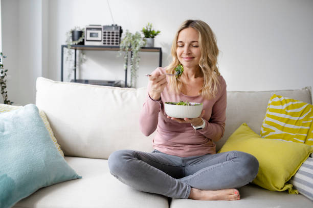 smiling woman with bowl of salad sitting on sofa - food stock pictures, royalty-free photos & images