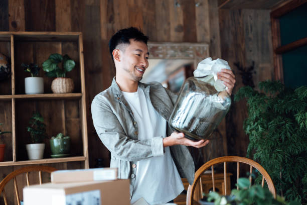 smiling young asian man looking happy as he unboxing the delivered package from online purchases at home, take out a glass vase. online shopping, enjoyable customer shopping experience - home decoration stock pictures, royalt