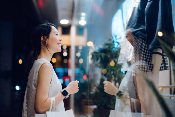 smiling young asian woman carrying a paper shopping bag, standing outside a boutique looking at shop window in the evening in city - fashion stock pictures, royalty-free photos & images