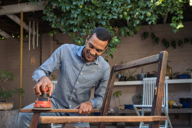 smiling young man sanding a chair outdoors - home decoration stock pictures, royalty-free photos & images