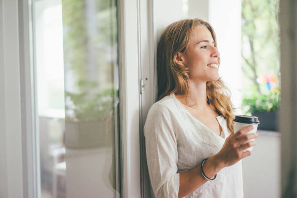 smiling young woman holding takeaway coffee at the window - junk food stock pictures, royalty-free photos & images