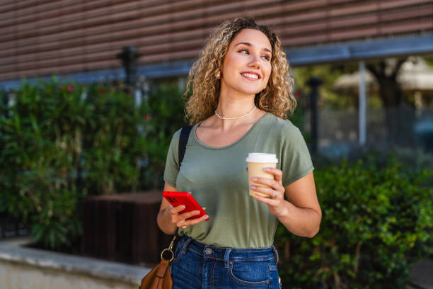smiling young woman walking with coffee and smartphone in city - junk food stock pictures, royalty-free photos & images