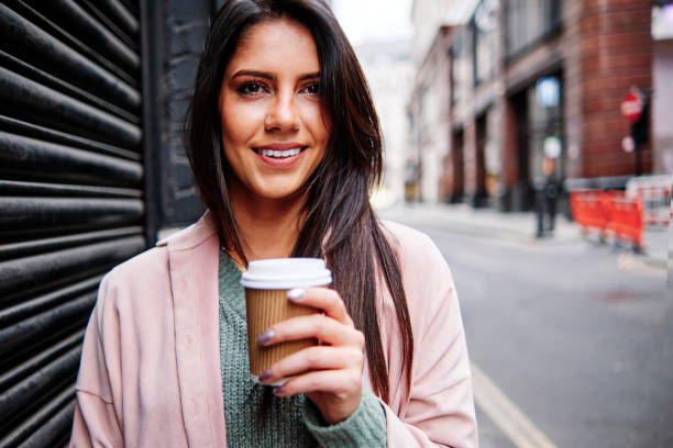 smiling young woman with disposable coffee cup standing by shutter in city - junk food stock pictures, royalty-free photos & images