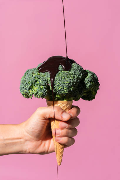 studio shot of man's hand holding ice cream cone with broccoli on top and pouring chocolate sauce, against pink background - food stock pictures, royalty-free photos & images