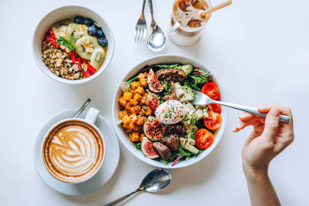 table top view of a woman eating healthy beef cobb salad bowl with greens, avocado, cherry tomatoes, figs, pumpkin and soft boiled egg, vegan chia and quinoa granola fruit bowl and coffee by the side. healthy food trend. nutr