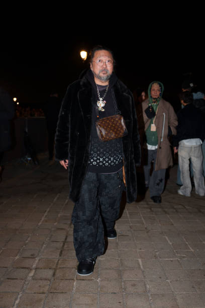 Takashi Murakami wears glasses, a black fluffy coat, a necklace depicting a cartoon character, a black pullover with silver shiny Louis Vuitton...