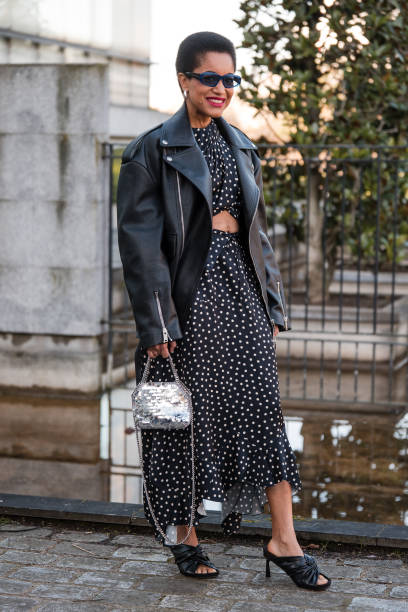 Tamu Mcpherson wears black and white polka dot dress, black leather jacket, blue sunglasses, silver Stella McCartney bag, black sandals, outside...