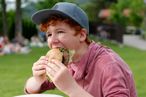teenage boy with baseball cap eating hamburger - junk food stock pictures, royalty-free photos & images